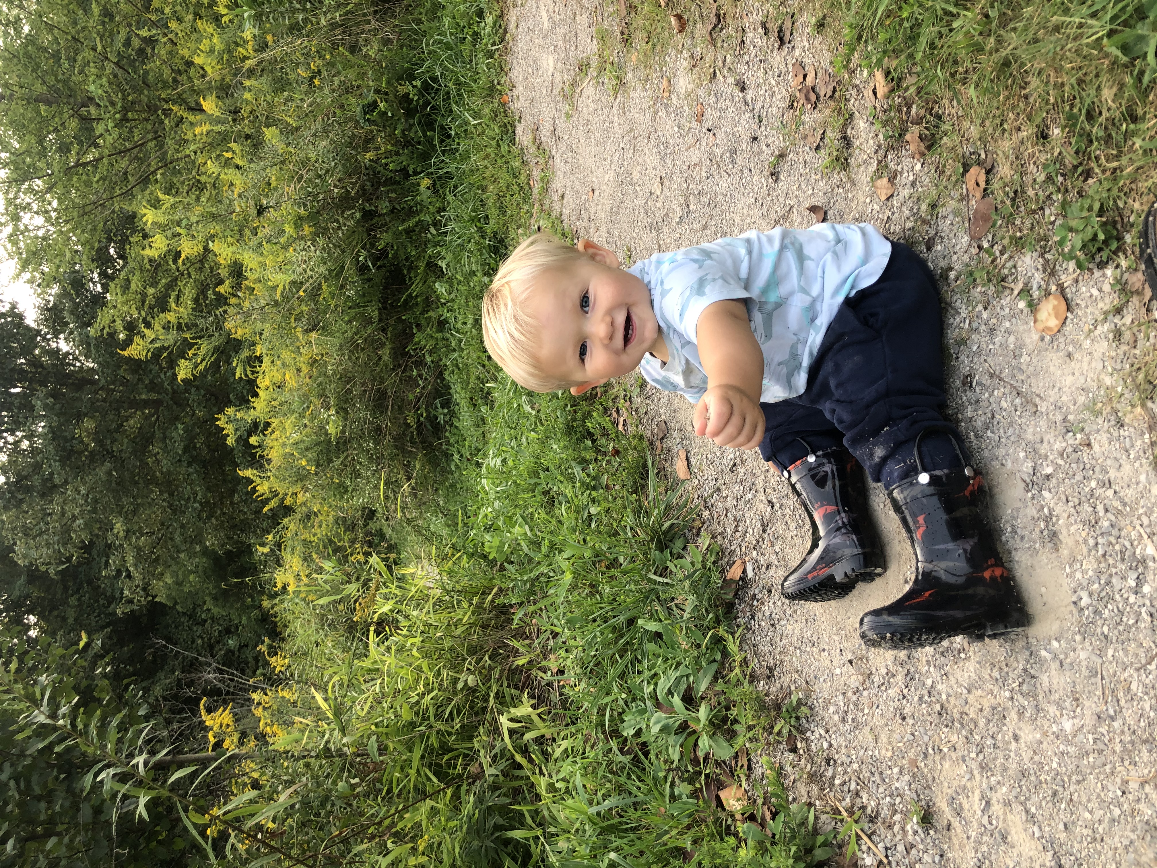 A toddler boy sits on a trail and smiles at the camera